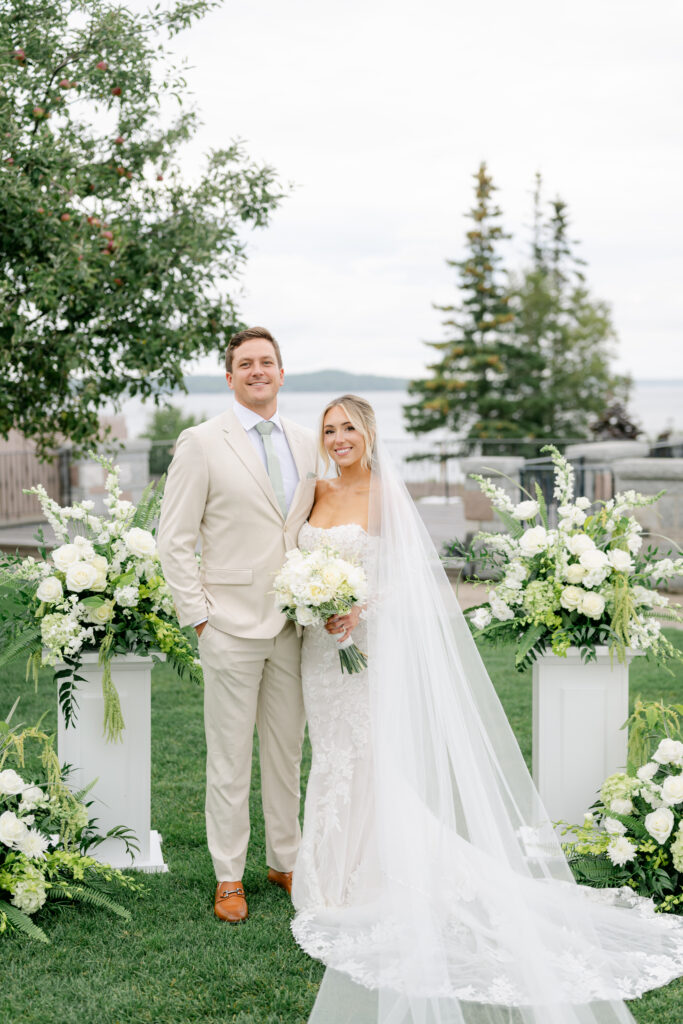 Wedding couple standing together at their Bar Harbor Club wedding ceremony with lush white floral arrangements and the Maine coastline behind them.