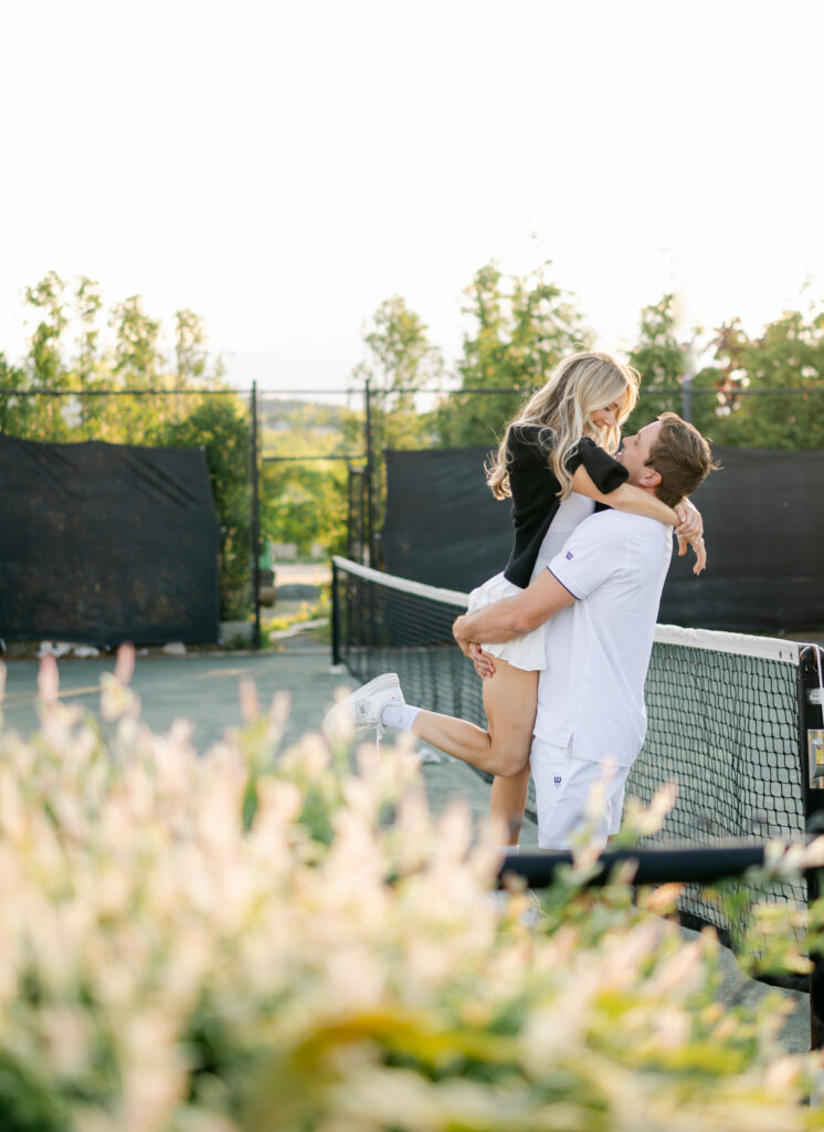 Bride-to-be lifted into her partner’s arms on an outdoor tennis court at Bar Harbor Club, photographed by a Maine Engagement Photographer and Maine Wedding Photographer in soft evening light.