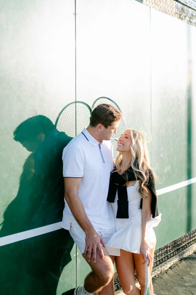 Couple dressed in white leaning against a green tennis court wall at Bar Harbor Club, captured by a Maine Wedding Photographer specializing in Maine engagement photography.